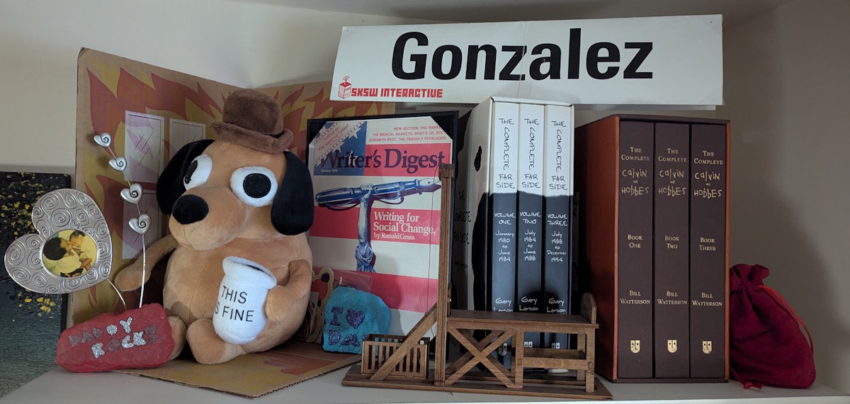"This is Fine" stuffed dog; a framed Writer's Digest cover; collected editions of The Far Side and Calvin and Hobbes. In front, a miniature guillotine.
