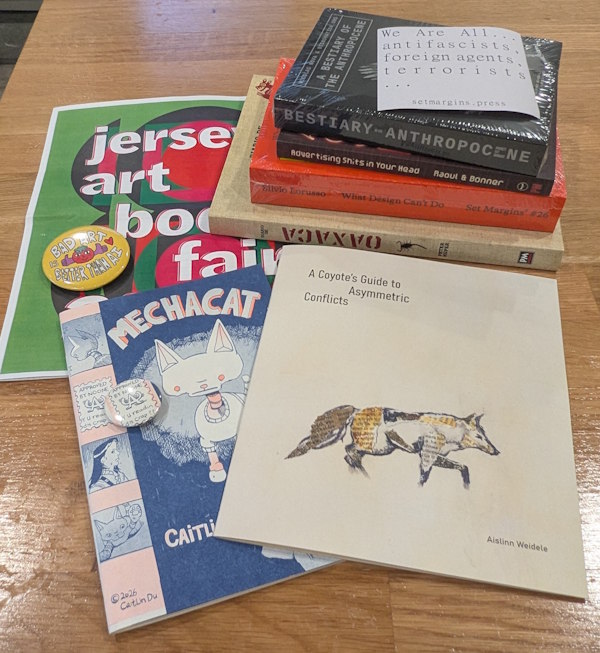 A stack of books and graphic novels, with a comic, a zine, two two art buttons, and a poster for the Jersey Art Book Fair.